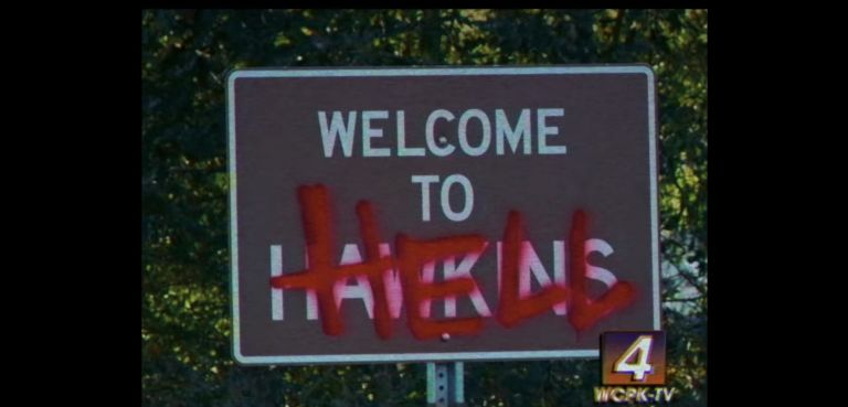 Una señal de tráfico que dice "Bienvenido a Hawkins" con la palabra "Infierno" pintada con aerosol sobre la palabra "Hawkins" en una escena de la temporada 3 de "Stranger Things"